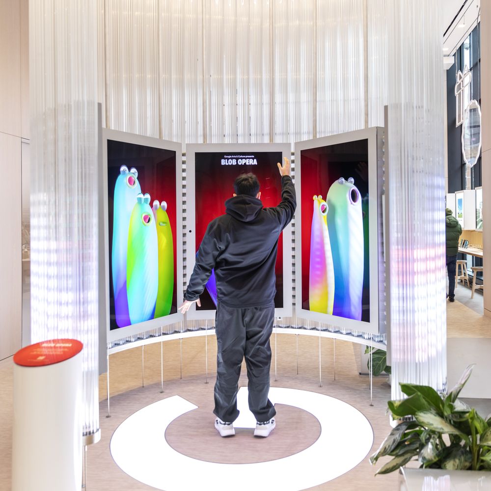 Guest standing inside the Blob Opera installation with arms raised, controlling colorful singing blobs across three vertical screens