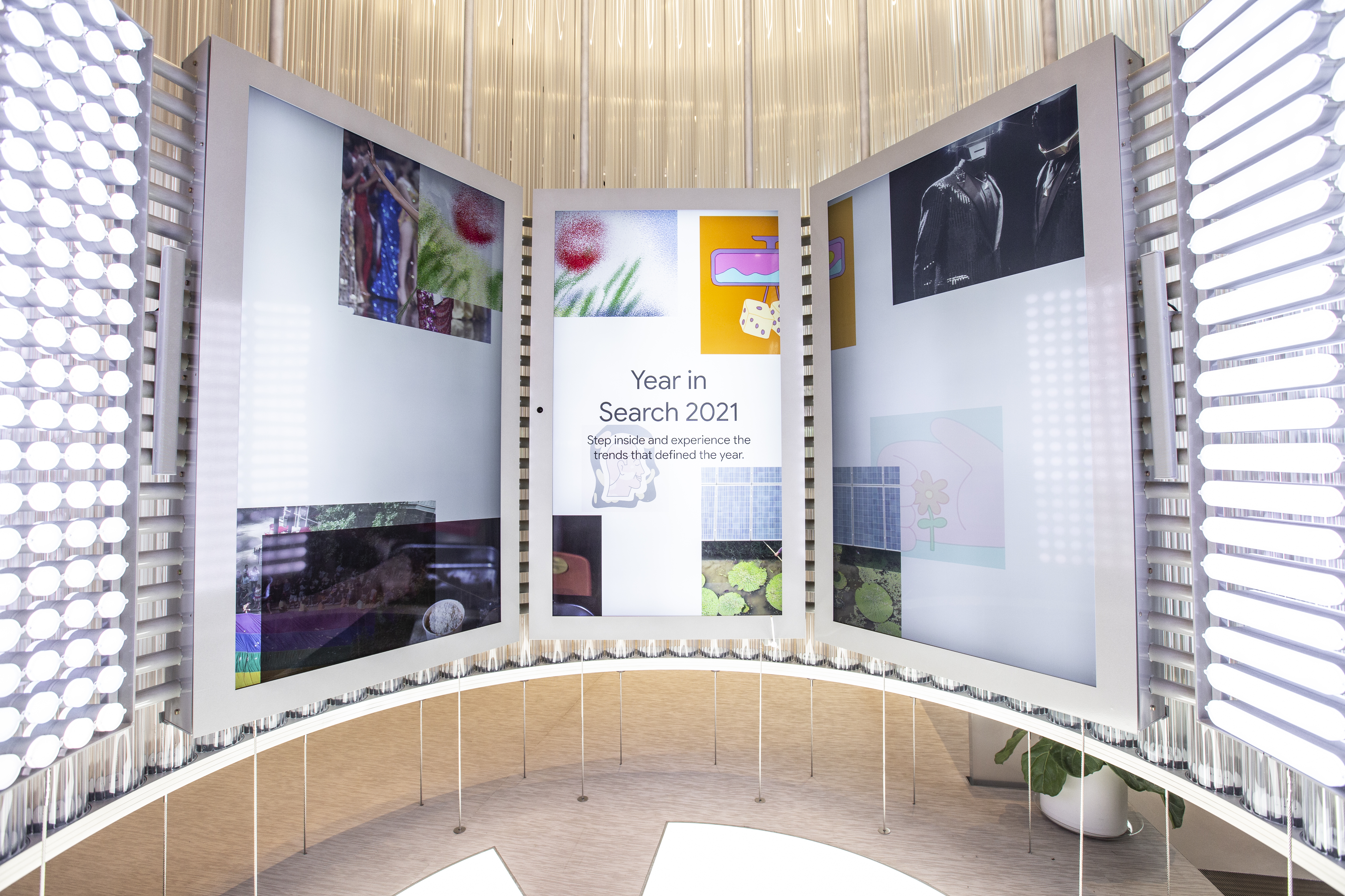 Close-up of the curved video-wall inside the Imagination Space — three tall vertical screens in the ambient state, the center reading &ldquo;Year in Search 2021 — Step inside and experience the trends that defined the year&rdquo; with collage tiles of the year's defining moments arranged around it; flanking white perforated columns with halo lighting frame the scene, and the hanging fiber strands of the dome canopy run overhead