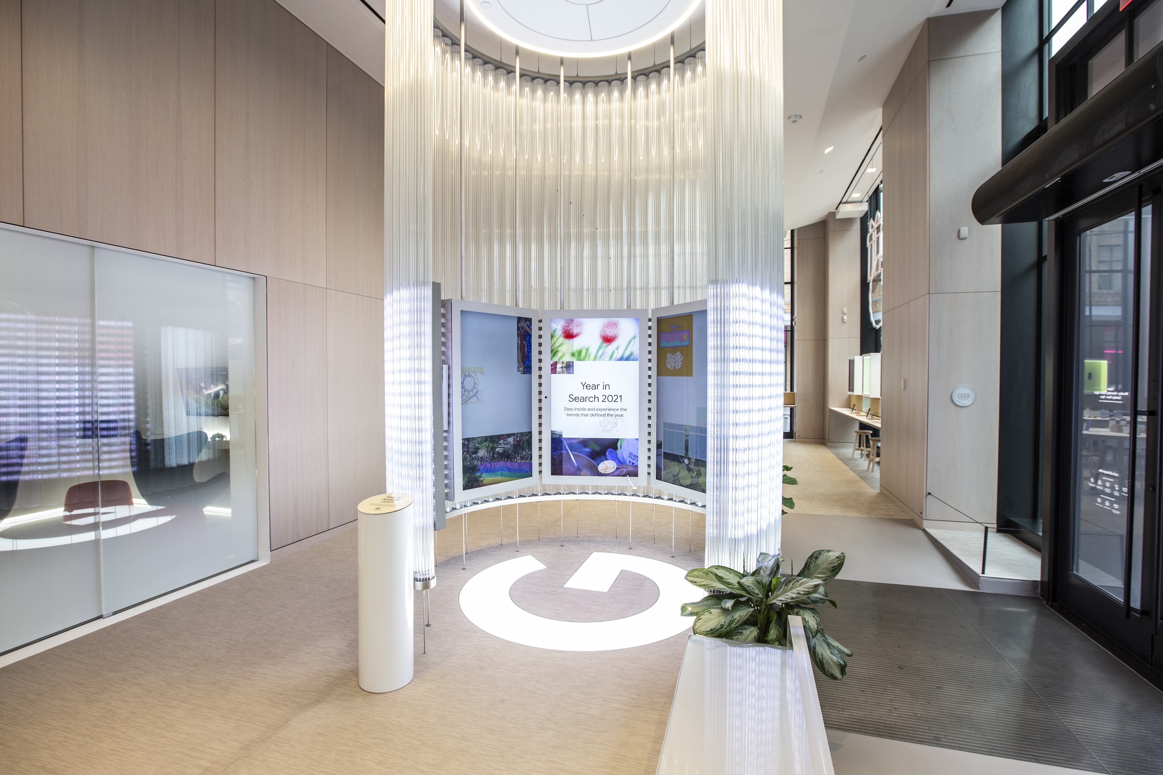 Year in Search exhibit in its ambient state inside the Google Store — the 17-foot circular glass structure lit from within with hanging fiber-optic strands, framing a curved video wall that reads &ldquo;Year in Search 2021 — Step inside and experience the trends that defined the year&rdquo; over a collage of moments; a large white Google G is set into the floor at the entrance, with two illuminated activation pillars flanking the doorway