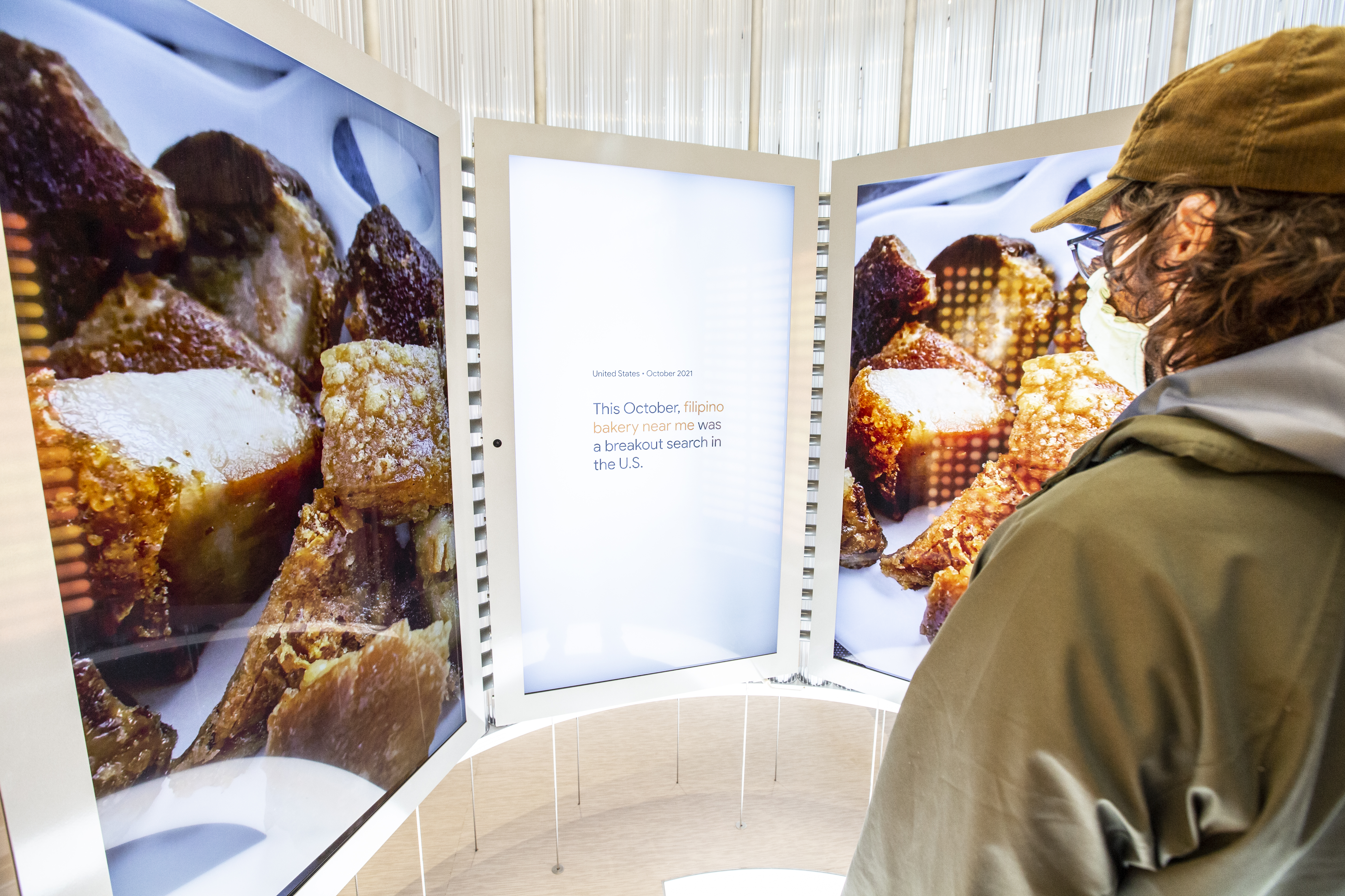 Deep-dive video playing on the curved video wall — the left and right screens show full-bleed close-ups of crispy lechon-style fried food, while the center screen reads &ldquo;United States · October 2021 — This October, filipino bakery near me was a breakout search in the U.S.&rdquo;; a guest in a tan corduroy cap watches from the right edge of frame
