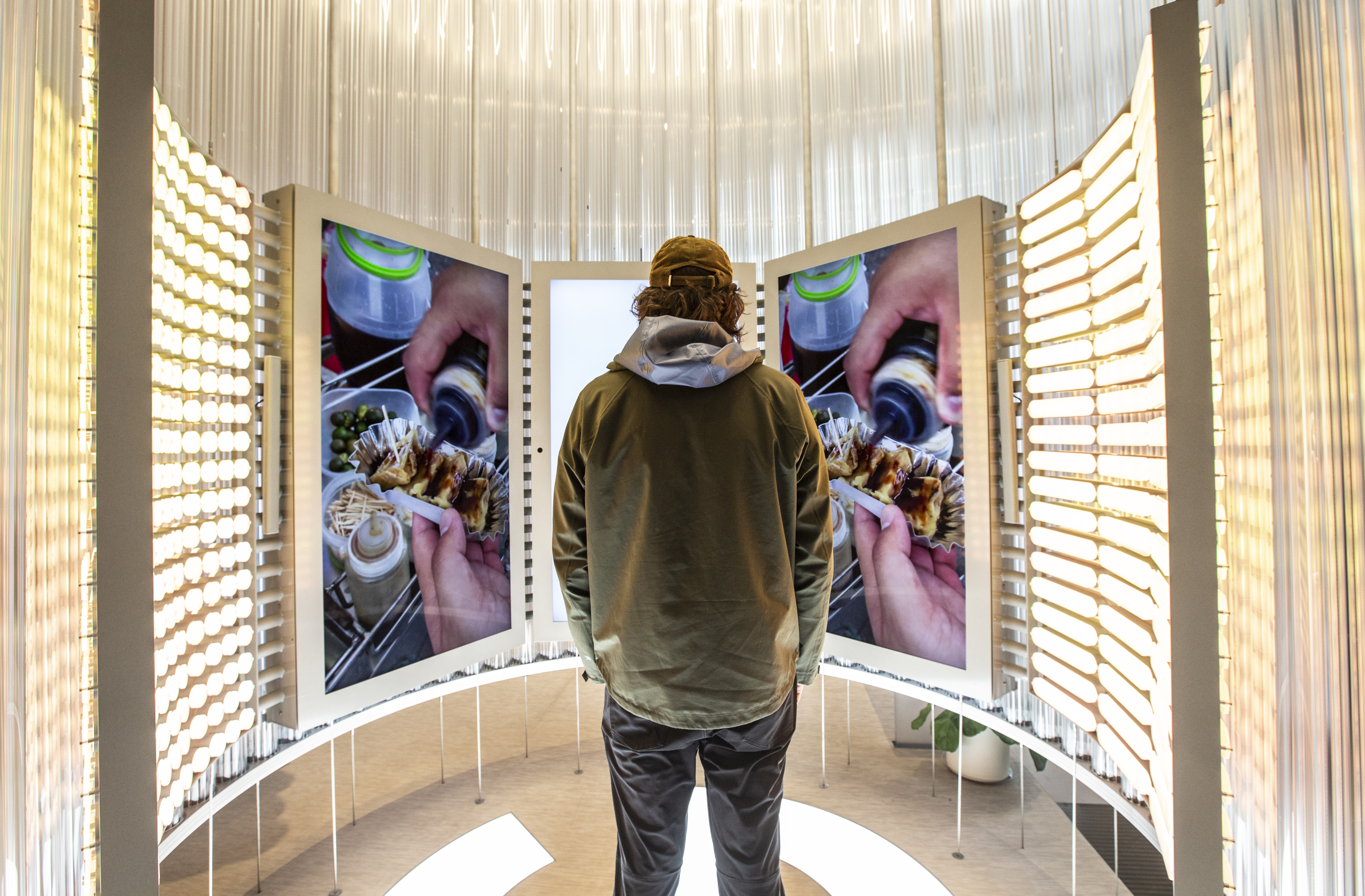 The same guest now stands centered on the Google G floor mark, watching the curved video wall from inside the Imagination Space — the left and right screens show a continuing deep-dive moment of someone pouring sauce over a tray of Filipino street food, while the activation columns flanking the screens glow warm and the fiber strands of the dome canopy fall around the scene
