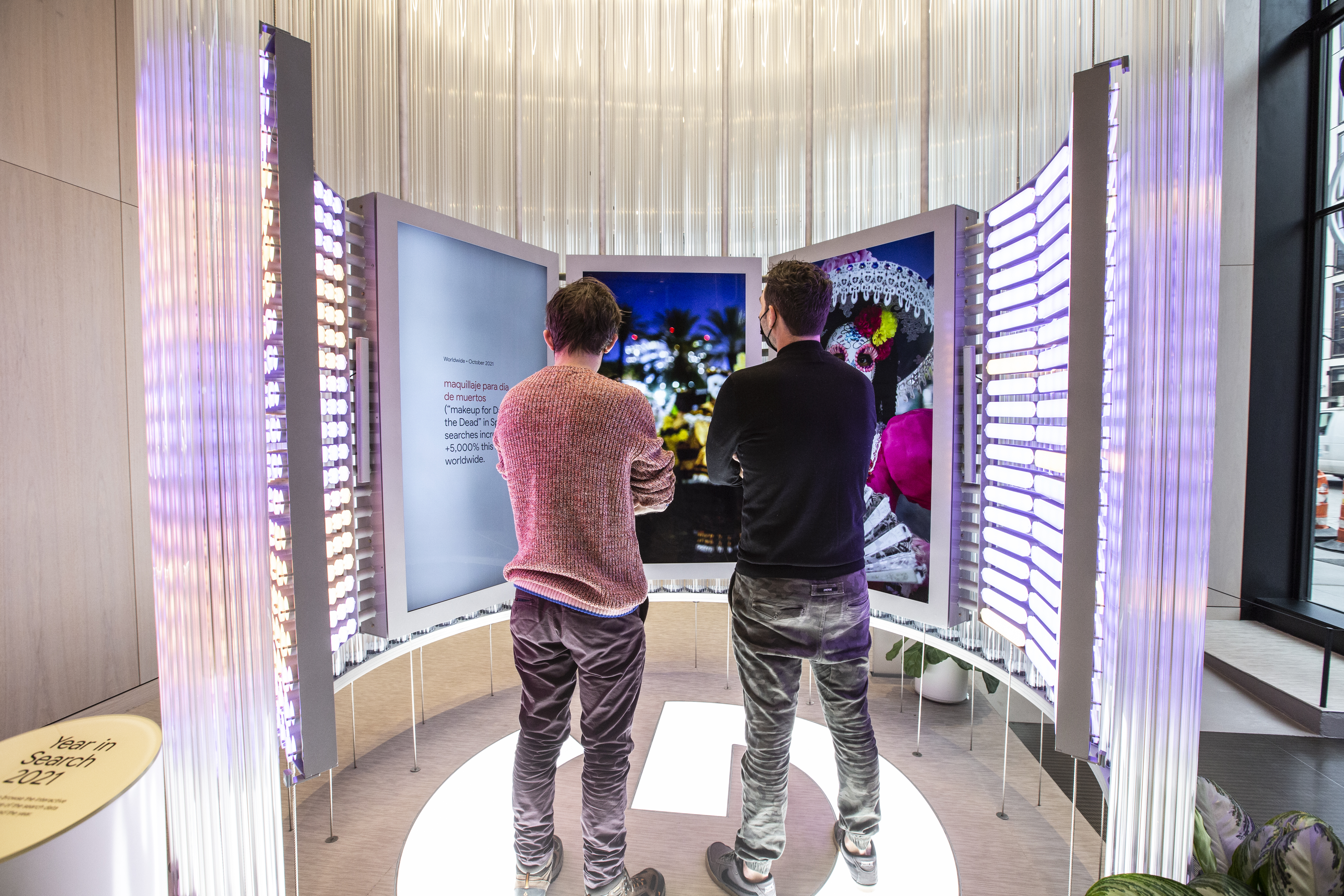 Two guests stand on the Google G floor mark inside the Imagination Space watching another deep-dive moment — the left screen reads &ldquo;Worldwide · October 2021 — maquillaje para dia de muertos (makeup for Day of the Dead in Spanish) searches increased +5,000% this worldwide&rdquo; in red and gray type, while the center and right screens show full-bleed photography of a Día de los Muertos celebration with a sugar-skull-painted face and marigolds; the activation columns flanking the wall glow magenta to match the moment