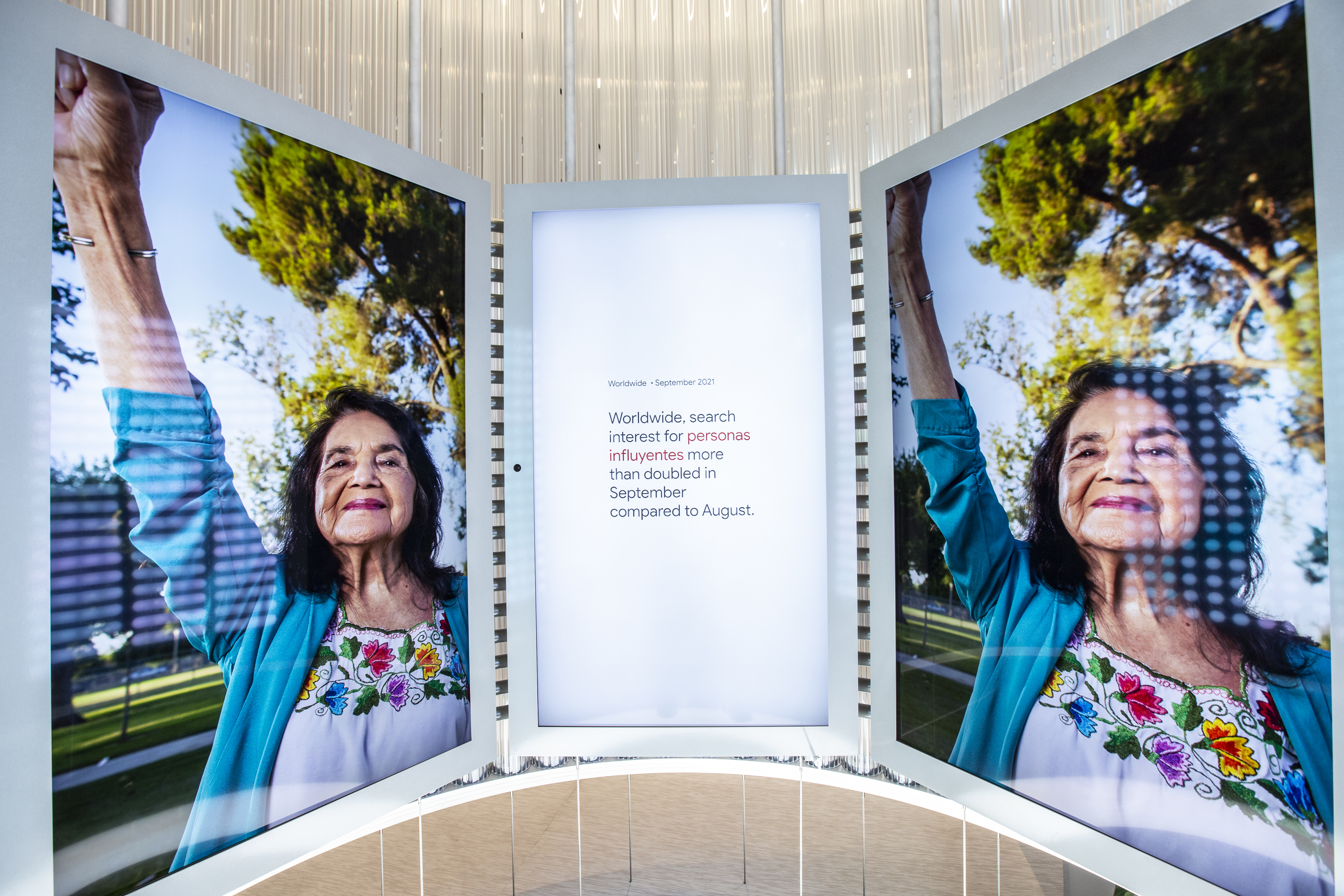 Another deep-dive on the curved video wall — the left and right screens show a full-bleed portrait of an older woman in a teal cardigan and embroidered floral blouse, fist raised triumphantly under a tree-lined sky, while the center screen reads &ldquo;Worldwide · September 2021 — Worldwide, search interest for personas influyentes more than doubled in September compared to August.&rdquo;