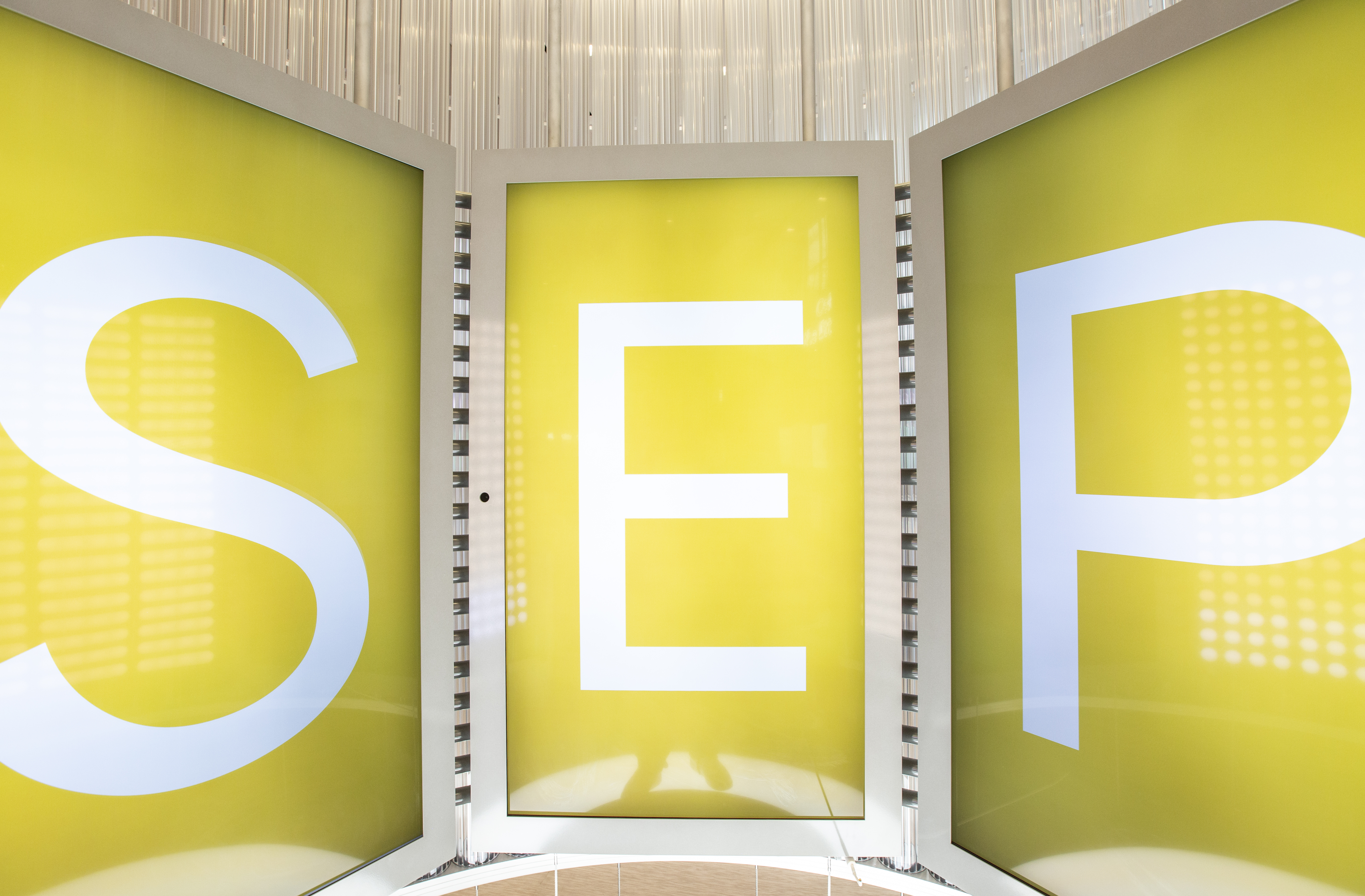 Tight-cropped view of the curved video wall mid deep-dive — the three vertical screens together spell out S-E-P in oversized white type on a chartreuse-yellow background, the month title card that opens each deep-dive segment