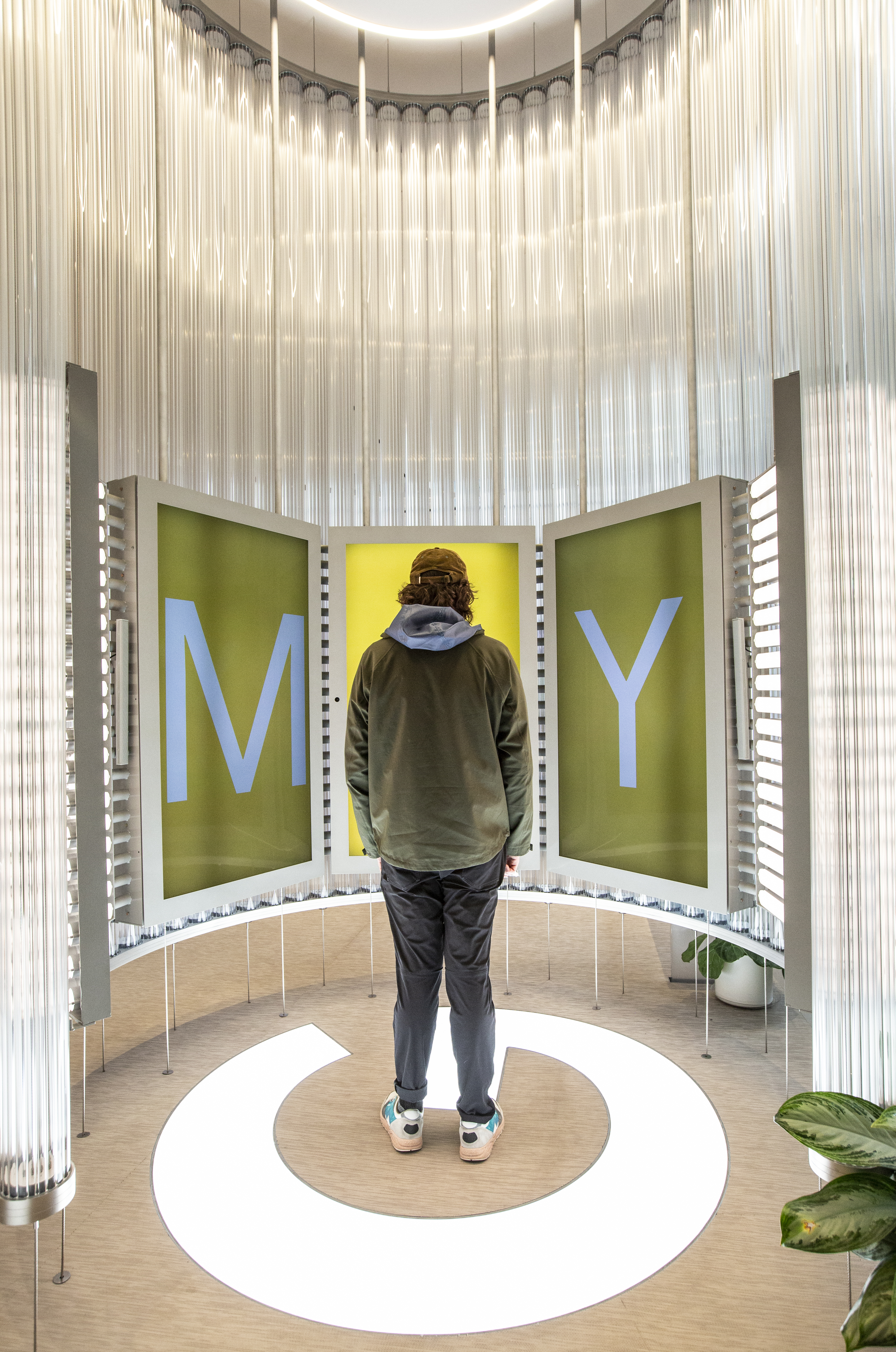 A guest stands on the white Google G floor mark inside the Imagination Space, facing the curved video wall — the three vertical screens have transitioned to a chartreuse-yellow background with the letters M, A, Y in tall pale-blue type, with the guest's body framed against the central A