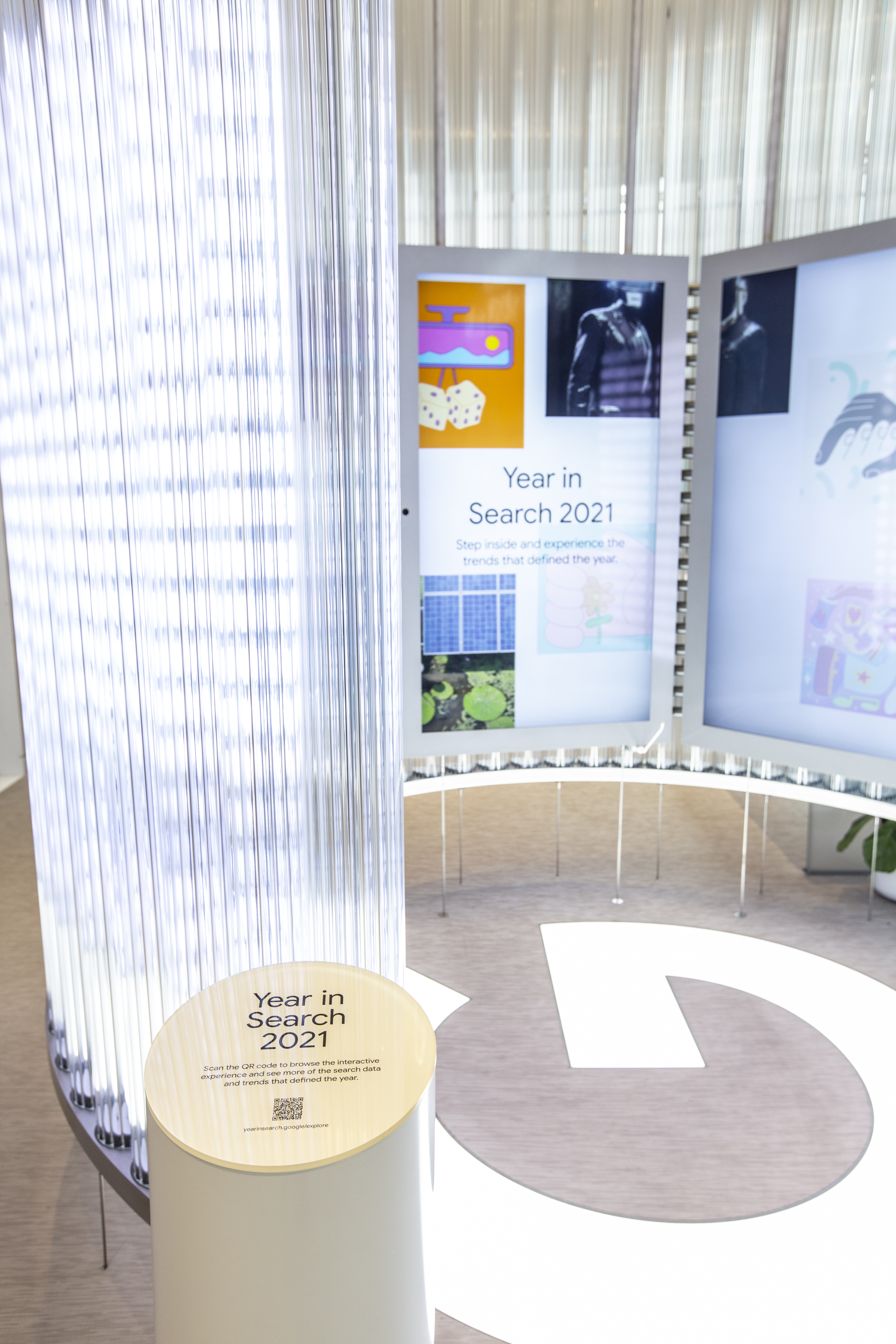 Close-up of the wooden disc placard atop one of the activation pedestals at the entrance to the Imagination Space — it reads &ldquo;Year in Search 2021 — Scan the QR code to browse the interactive experience and see more of the search data and trends that defined the year,&rdquo; with a QR code and the URL yearinsearch.google/explore beneath; behind it the curved video wall sits in its ambient state with collage tiles around the Year in Search 2021 invitation, and the Google G floor mark is visible at right