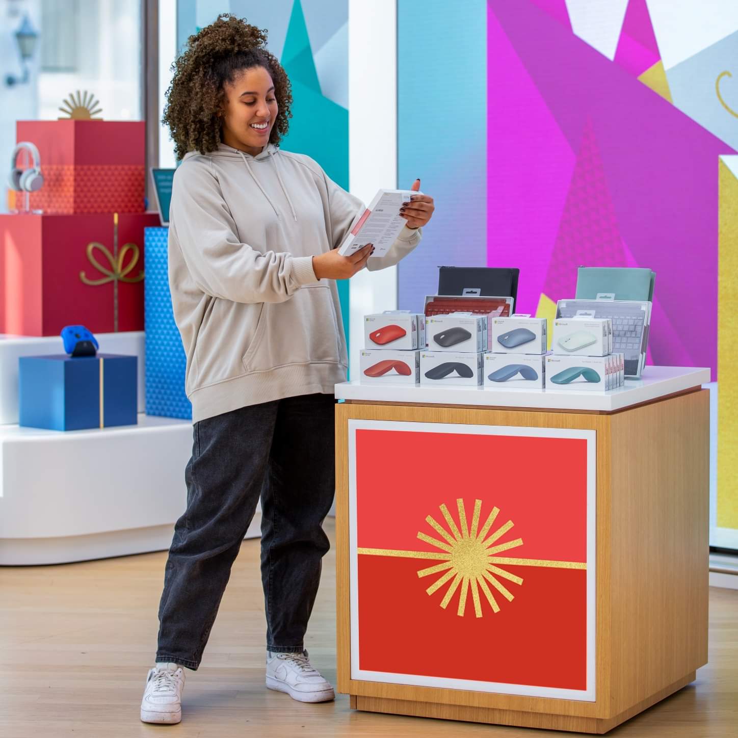 Guest browsing Microsoft accessories on a gift-wrapped display table inside a Microsoft store — wood and red gift-ribbon table topped with mice and keyboards, surrounded by oversized gift-box plinths in red and blue with a colorful holiday backdrop