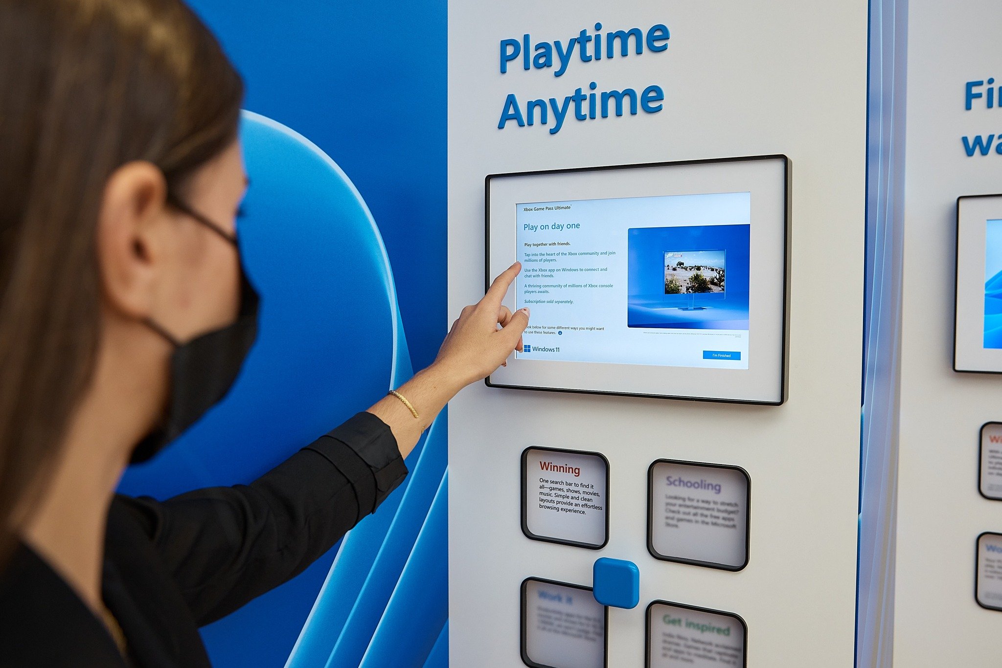 Close-up of one Windows 11 launch fin — a customer in a black mask reaches up to tap the Playtime Anytime Surface tablet showing the Xbox Game Pass Ultimate &ldquo;Play on day one&rdquo; screen, with four square tile cards labeled Winning, Schooling, Work it, and Get inspired mounted below and a small blue tactile selector between them