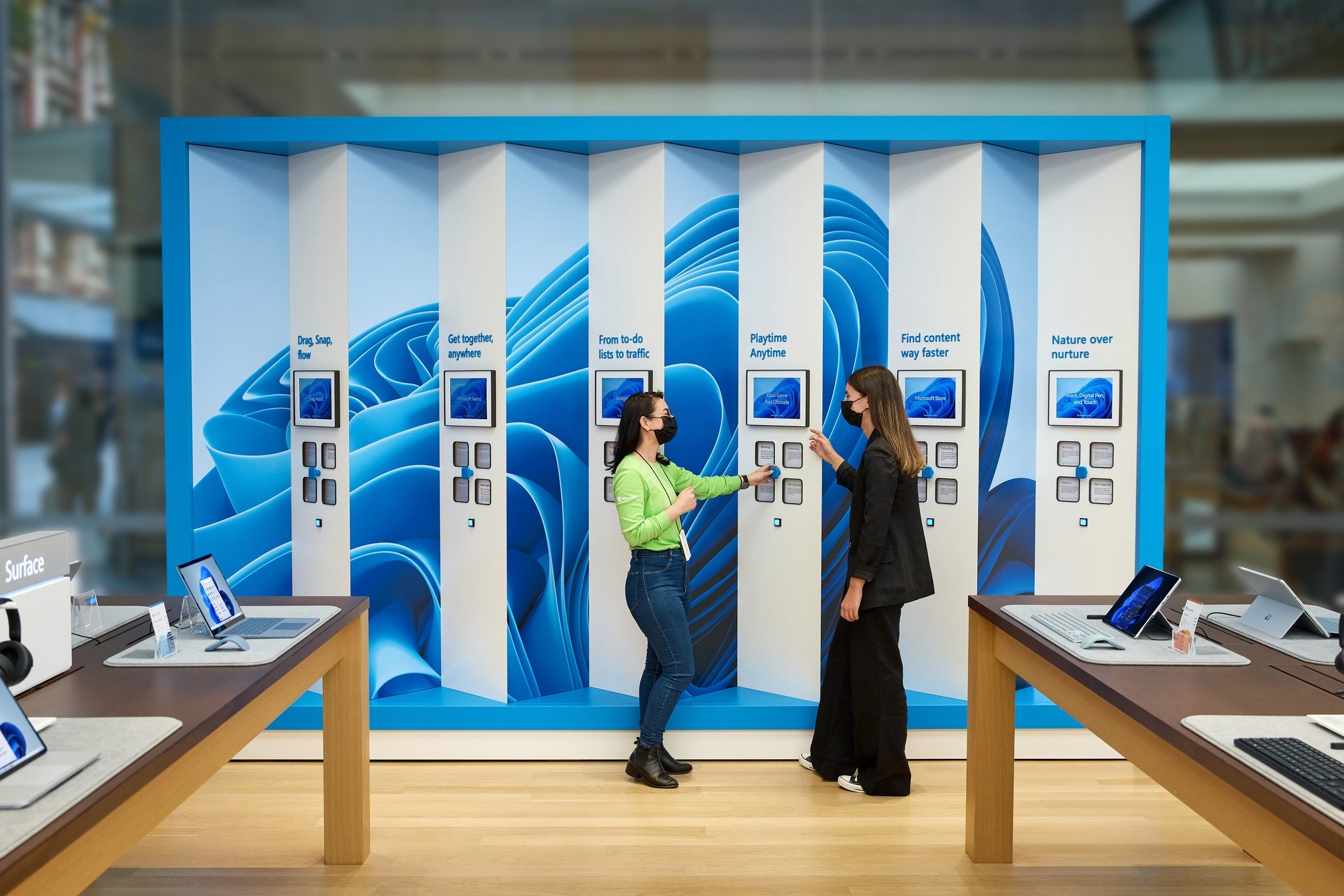 Final Windows 11 launch wall installed in the Sydney Microsoft store — six white fins set against the blue Windows 11 bloom backdrop, each fin holding a Surface tablet demoing a feature (Snap Assist, Microsoft Teams, Widgets, Xbox Game Pass, Microsoft Store, Voice/Pen/Touch) with a Microsoft associate guiding a customer through the interactives