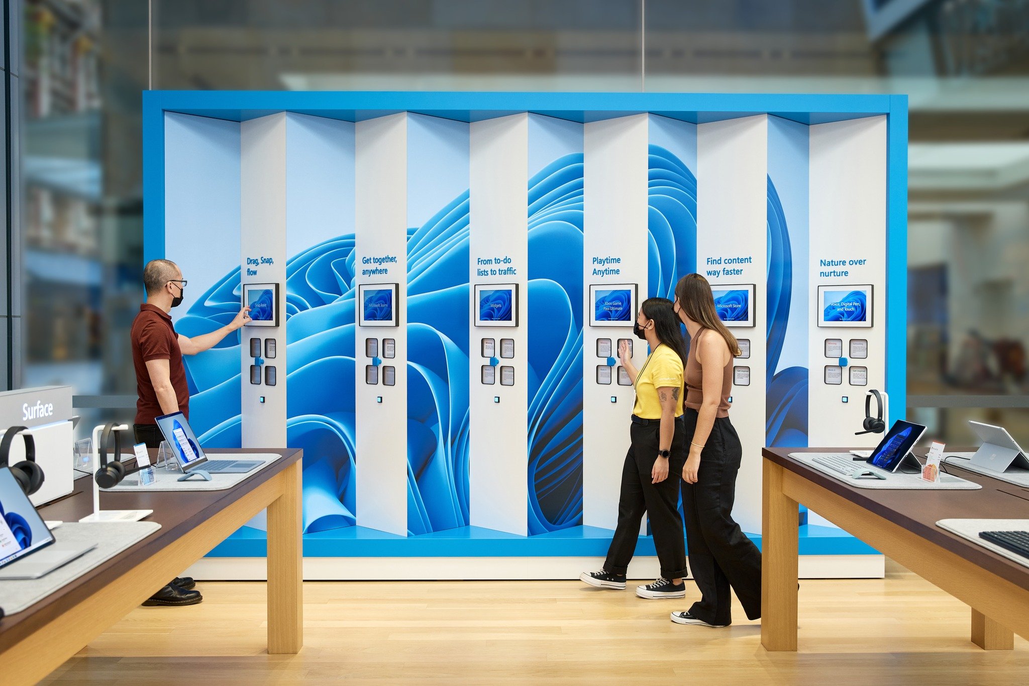Wide view of the Windows 11 launch wall in the Sydney Microsoft store — the full six-fin interactive wall with the blue bloom mural behind it, a customer in a maroon polo tapping the Snap Assist tablet on the left, and on the right a Microsoft associate in a yellow shirt walking another customer up to the Find content way faster fin; Surface devices on display tables flank the wall in the foreground