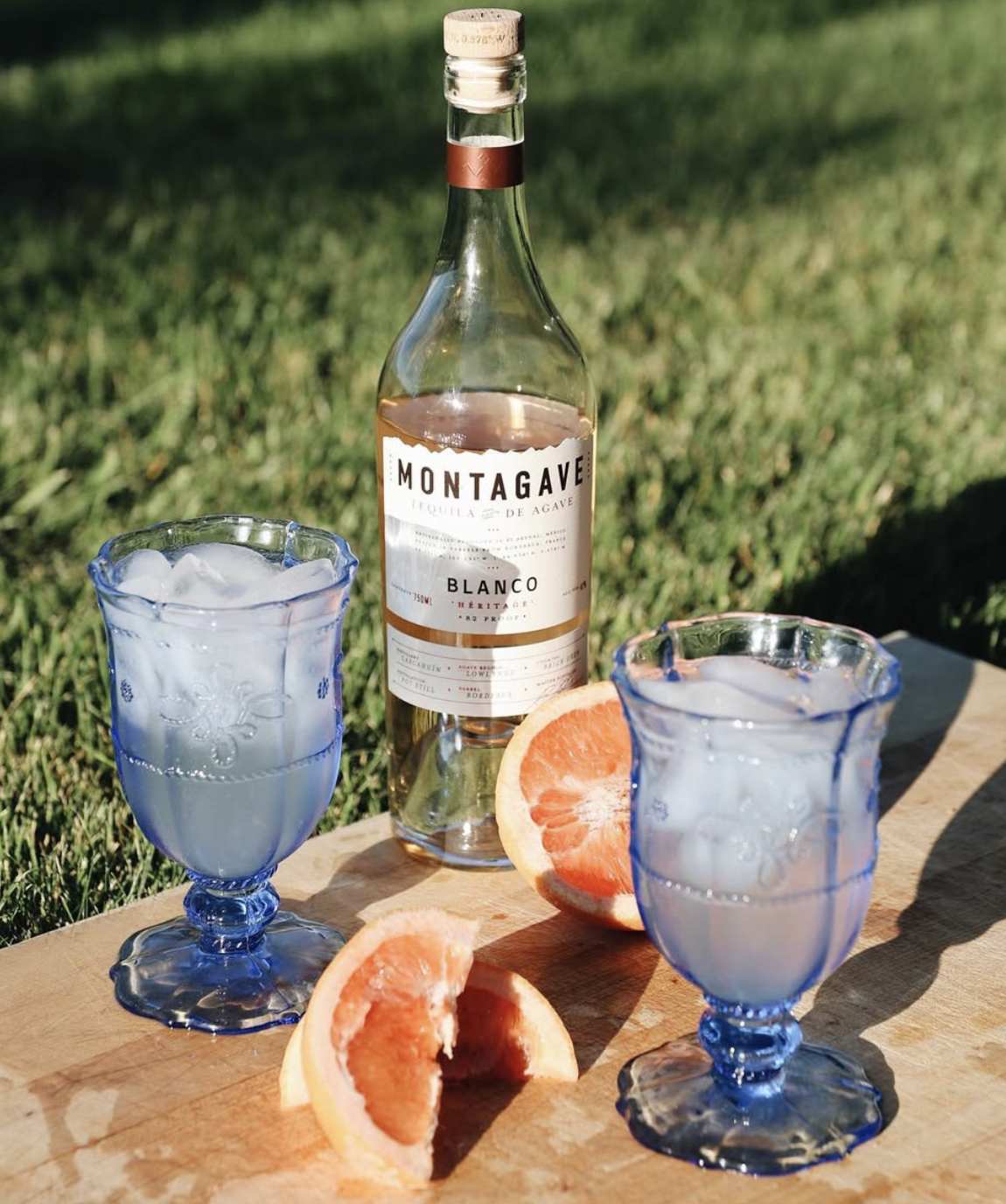 Bottle of Montagave Blanco tequila on a wood board outdoors, flanked by two ornate cobalt-blue goblets filled with ice and a clear cocktail, with a halved pink grapefruit and a fresh wedge resting beside the bottle and sunlit grass behind