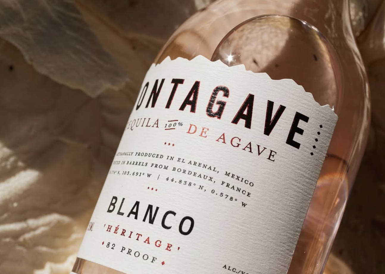 Tight close-up of the Montagave Blanco Héritage label on the curved glass bottle, showing the torn-edge cream paper, deckled black wordmark, red 100% De Agave callout, fine origin text, and the BLANCO 82 PROOF mark beneath, with diffused beige fabric in the background