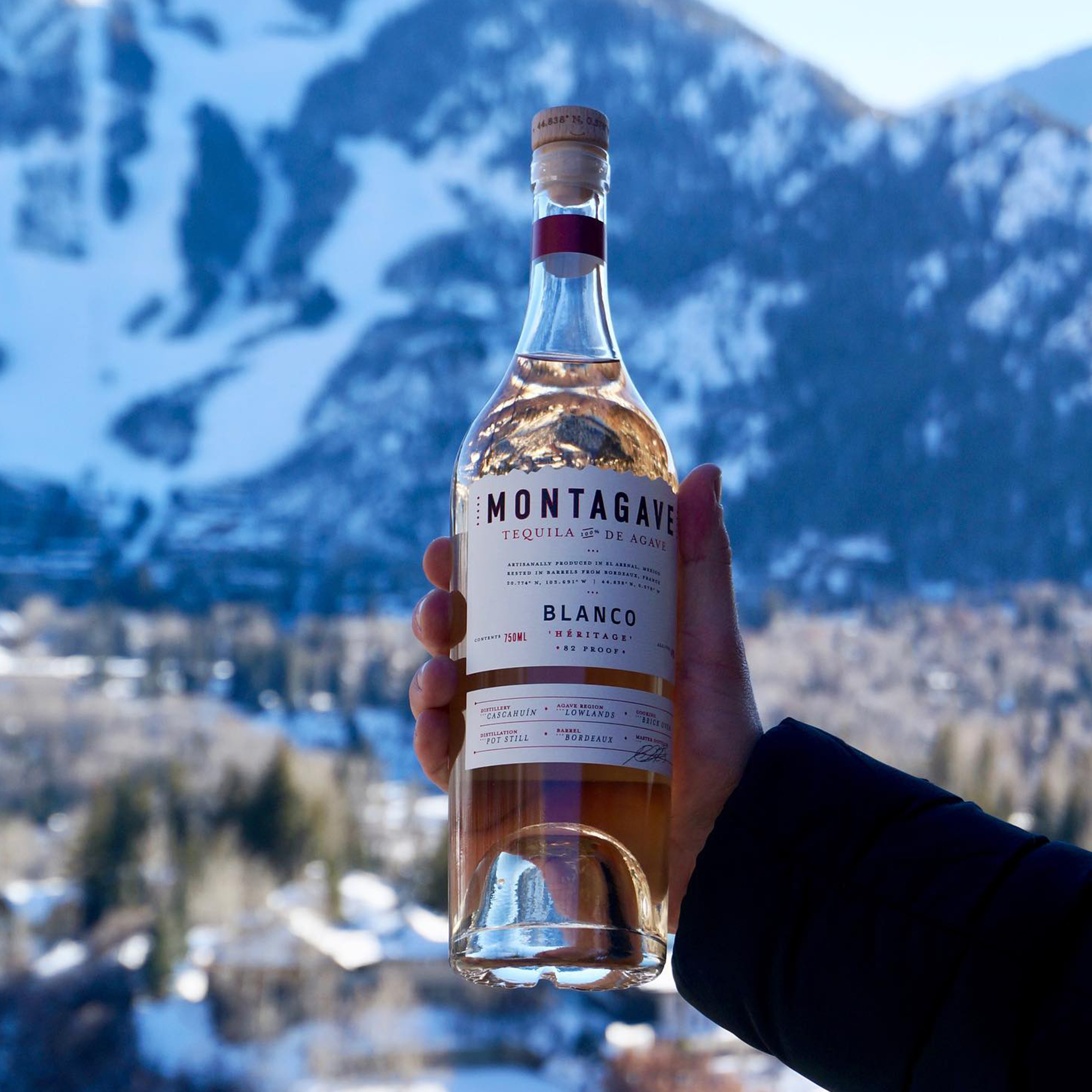 A hand holds up a clear glass bottle of Montagave Blanco Héritage tequila against a backdrop of snow-covered alpine peaks and a small village in the valley below, sunlight glinting off the bottle's torn-edge cream label