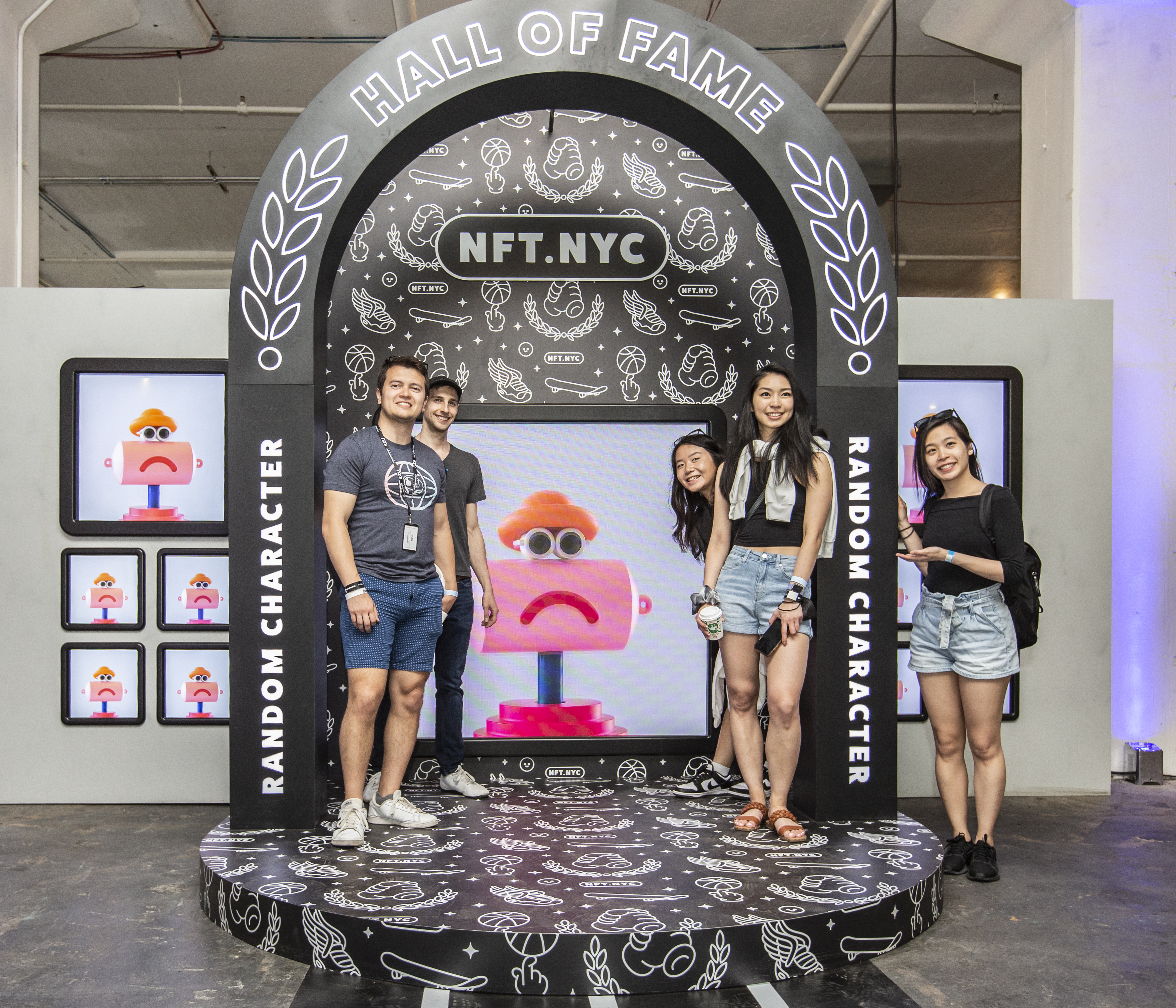 Four guests posing on the Hall of Fame photo-moment platform — a black NFT.NYC arch covered in line-art iconography frames a screen displaying a pink Random Character NFT, with a wall of character displays beside it