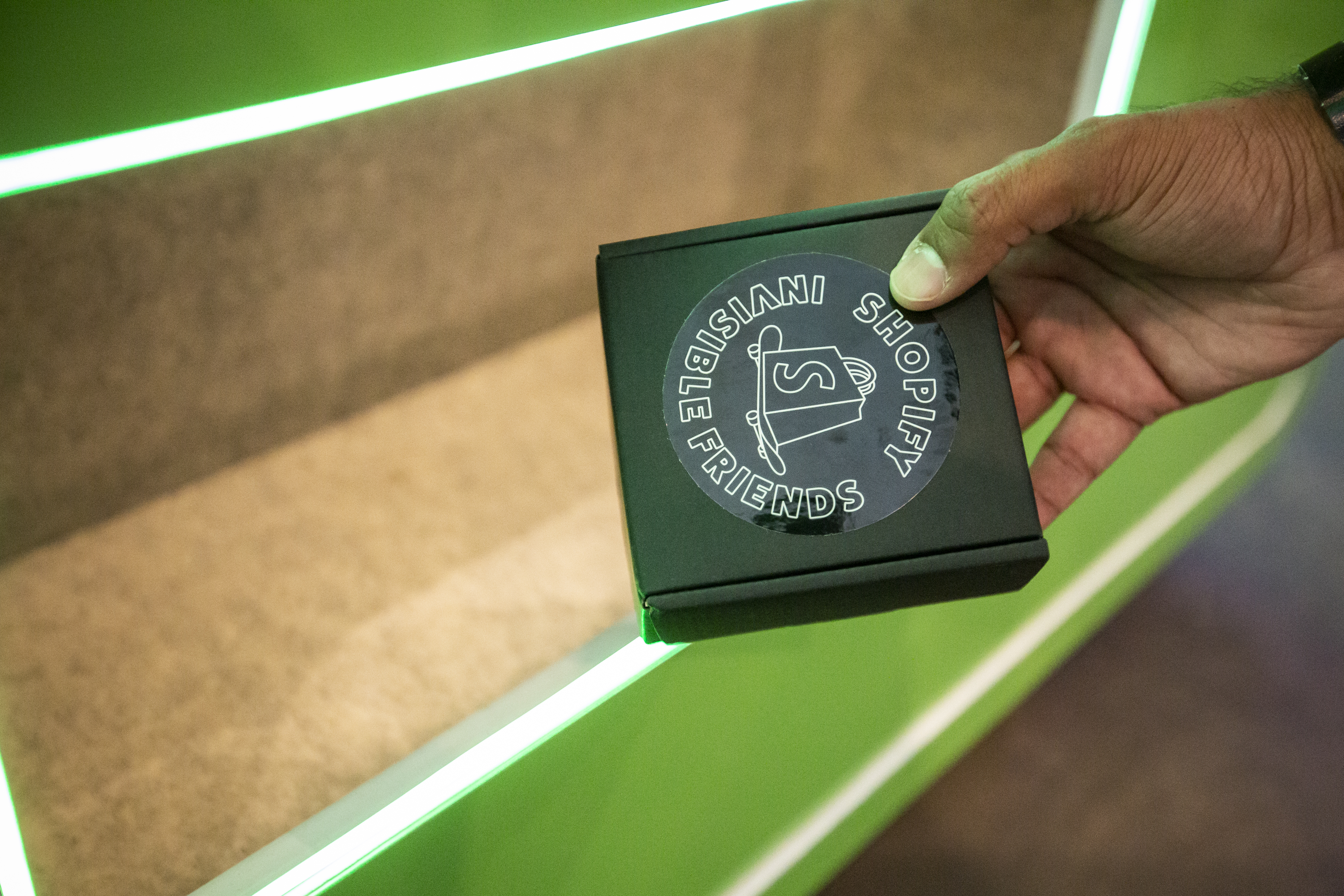 Hand holding the dispensed merch box — black box stamped with an Invisible Friends x Shopify circular sticker, framed by the green LED-lit interior of the vending machine