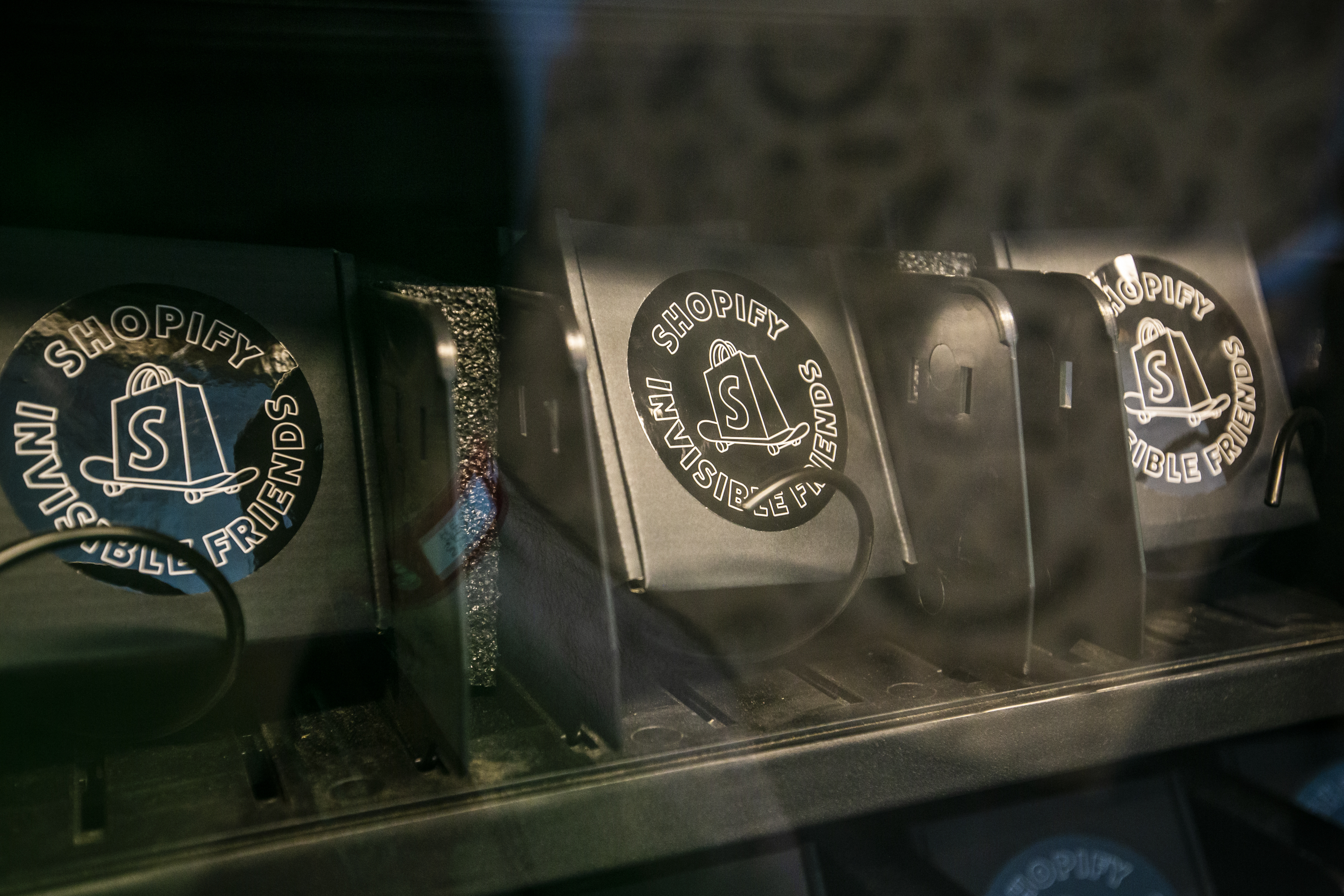 Close-up inside the vending machine — rows of black merch boxes stamped with circular Shopify x Invisible Friends stickers waiting on the dispenser coils behind the glass
