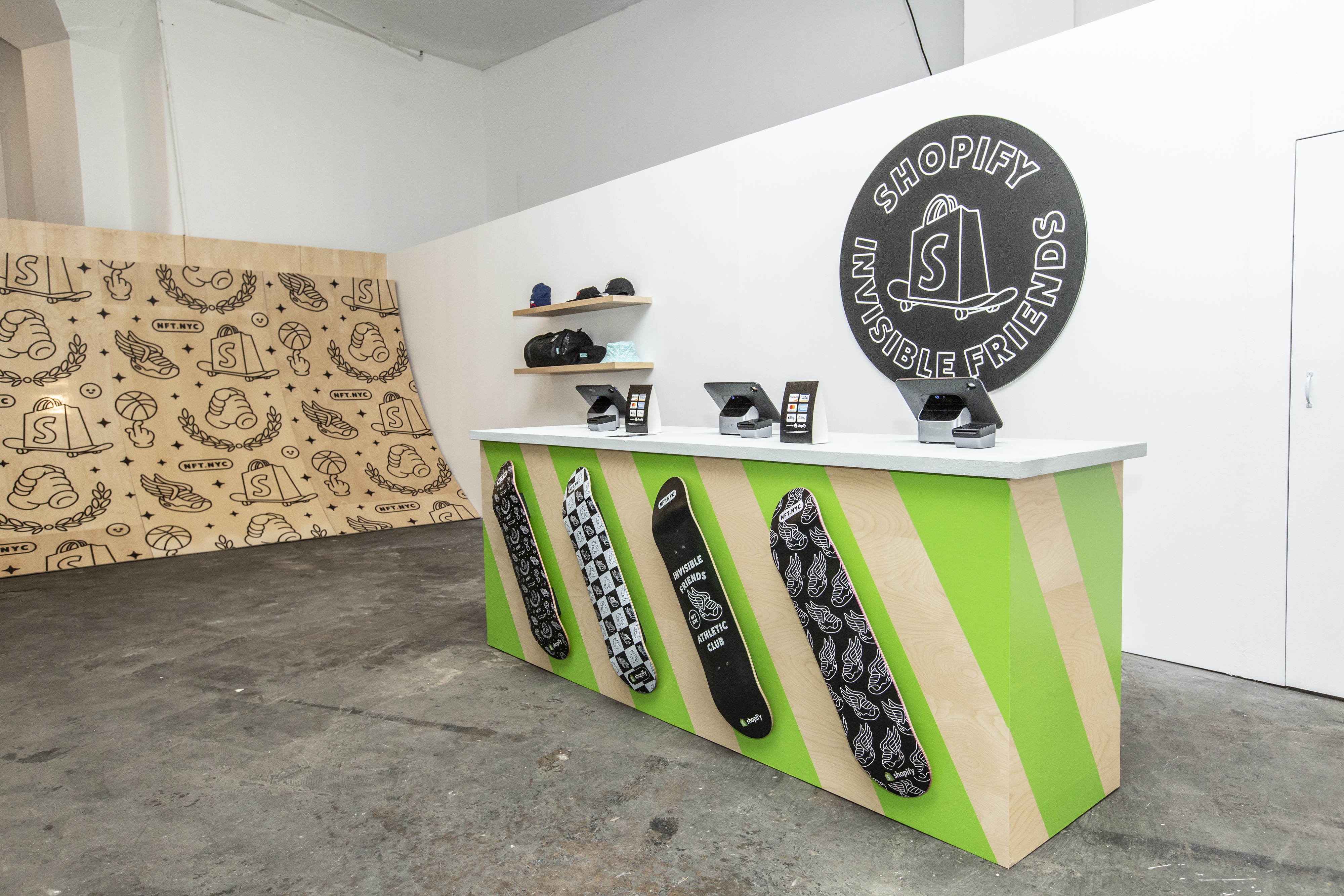 Merch station counter — green and wood striped front with four custom skateboard decks mounted to it, Shopify POS terminals on top, and a large Shopify x Invisible Friends circular logo on the back wall next to a pattern wall of NFT.NYC iconography