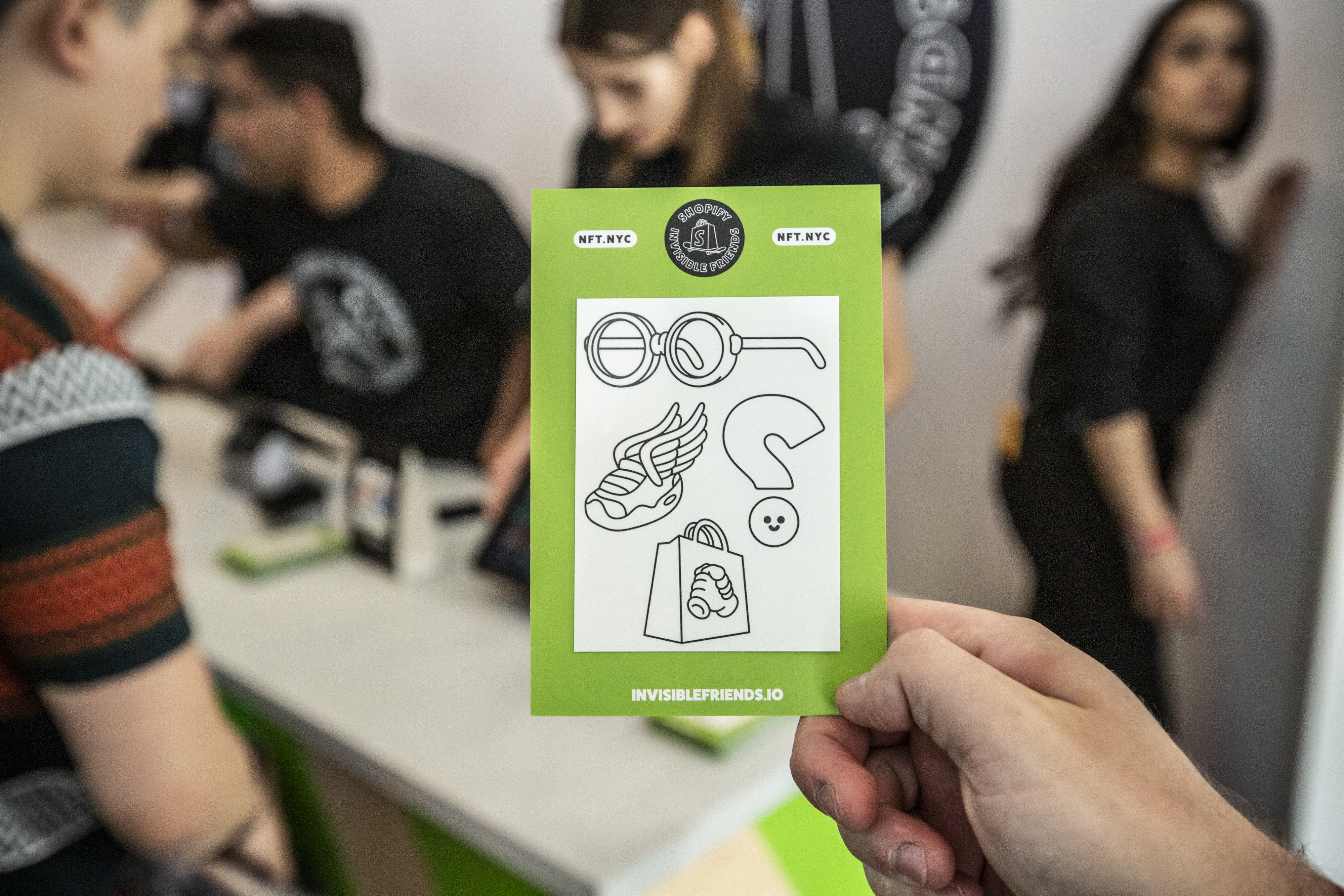 Hand holding a green Shopify x Invisible Friends sticker sheet — line-art icons of glasses, a winged sneaker, a Shopify shopping bag, and a smiley face, with NFT.NYC tags and an invisiblefriends.io URL