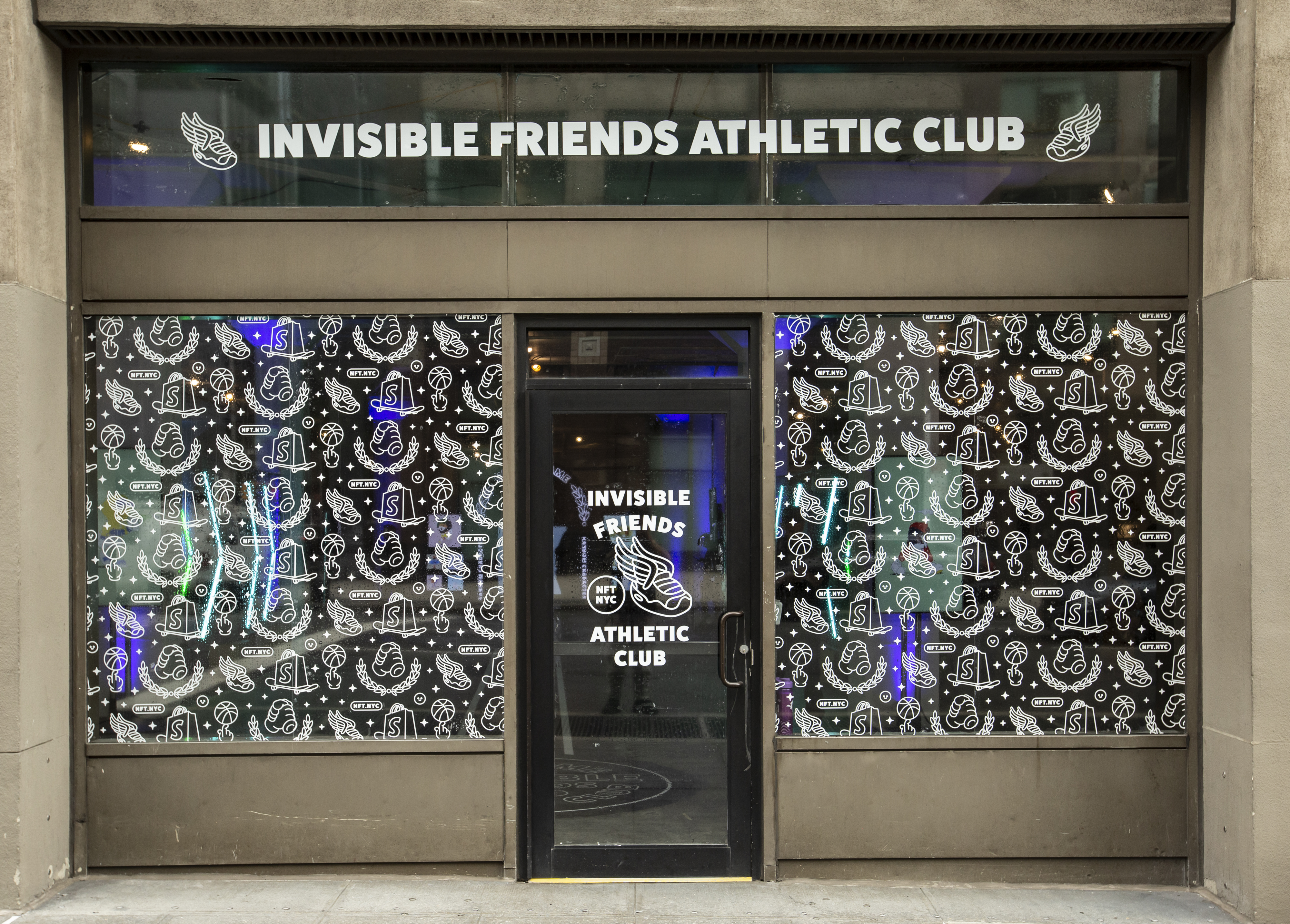 Storefront exterior of the activation — 'Invisible Friends Athletic Club' wordmark across the transom and patterned NFT.NYC iconography window decals on either side of a glass door with a winged sneaker logo