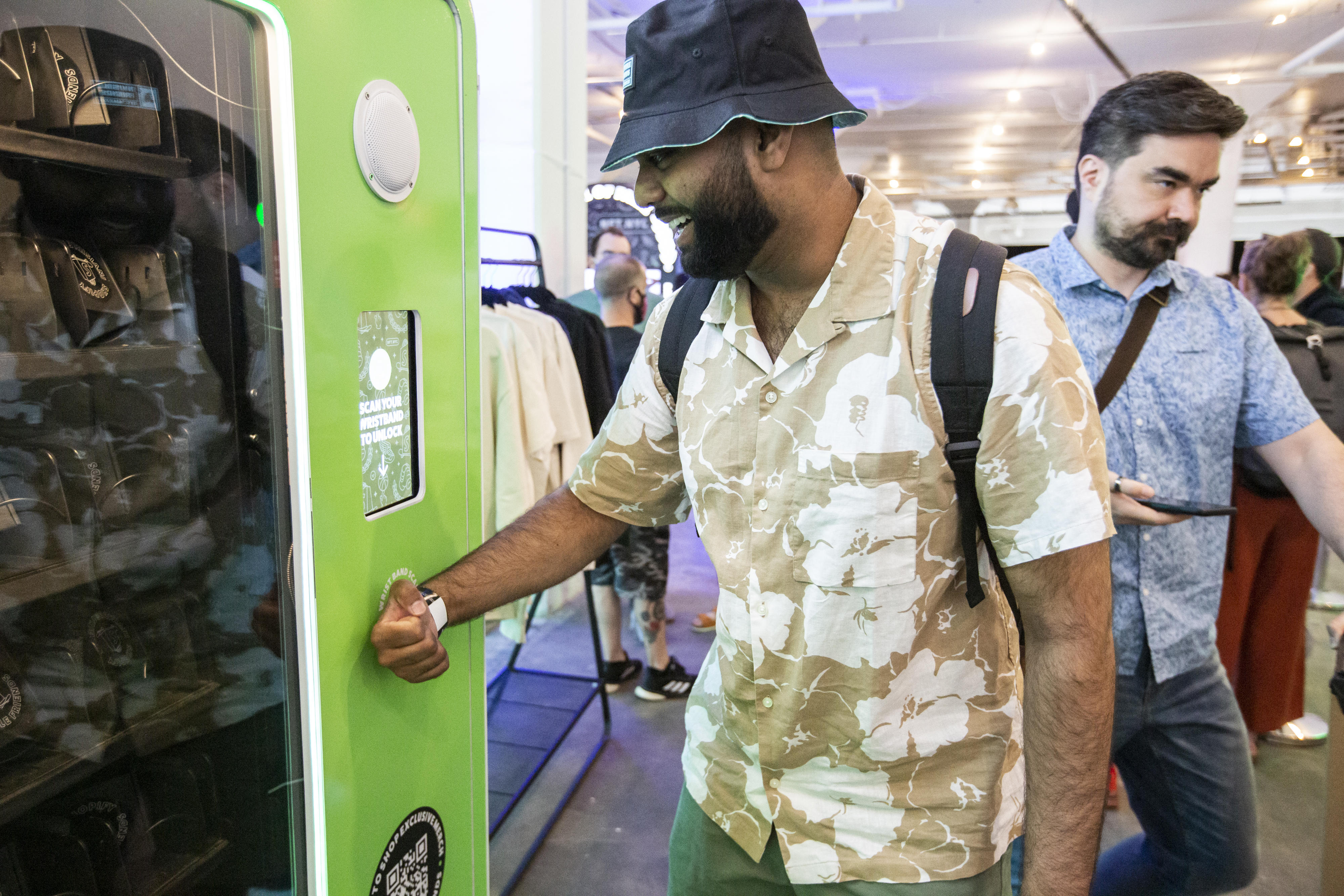 NFT holder scanning their wristband against the green Shopify-powered vending machine — on-screen prompt reads 'Scan your wristband to unlock' with apparel racks visible in the background